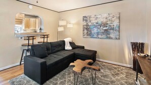 Una sala moderna con un sofá seccional, una mesa de centro con forma de guitarra, un ventilador de techo y una cocina visible a través de un arco.