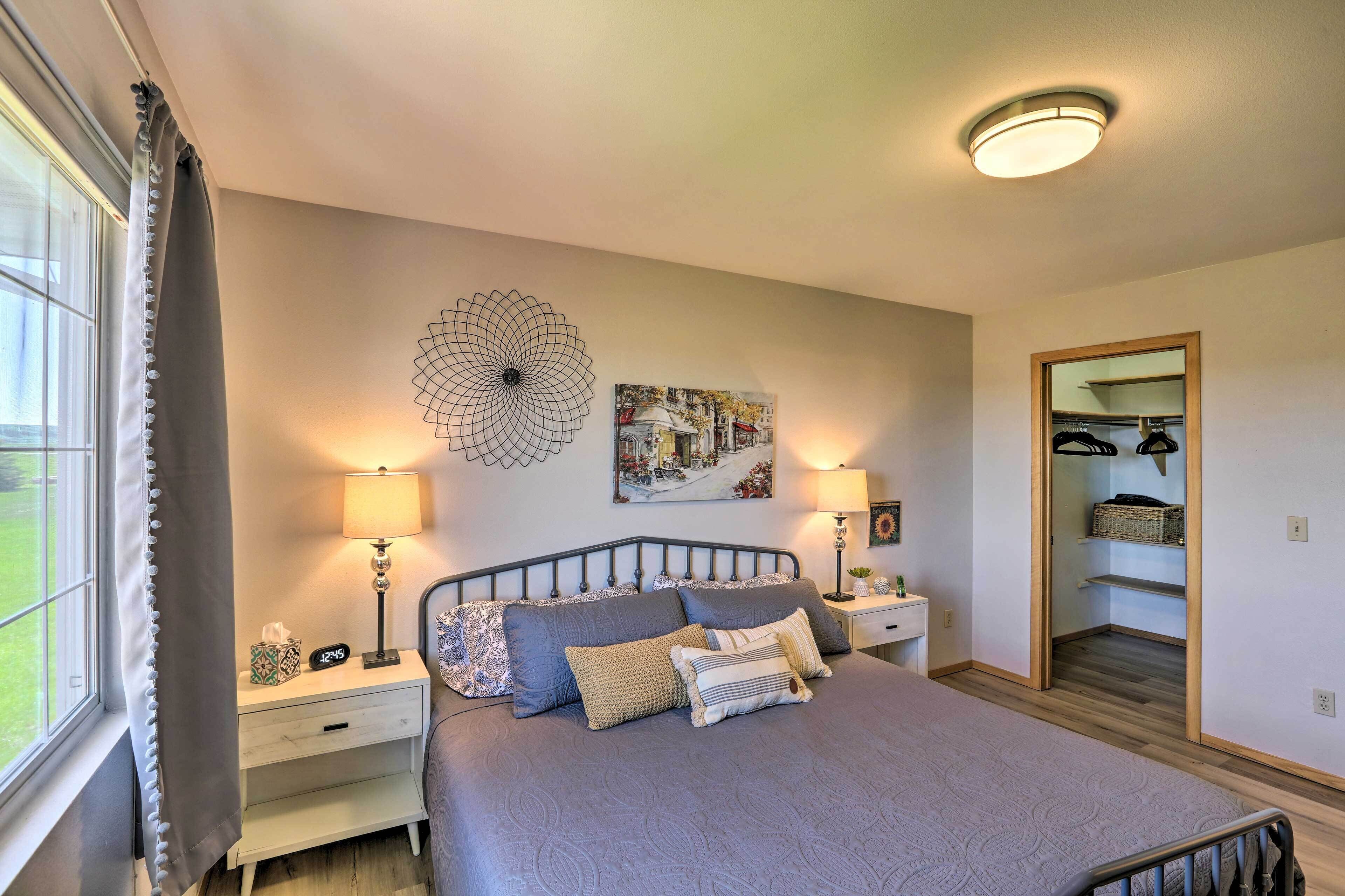 A bedroom with a bed, two bedside tables, a nightstand, a wall clock, a mirror, and a window with curtains.