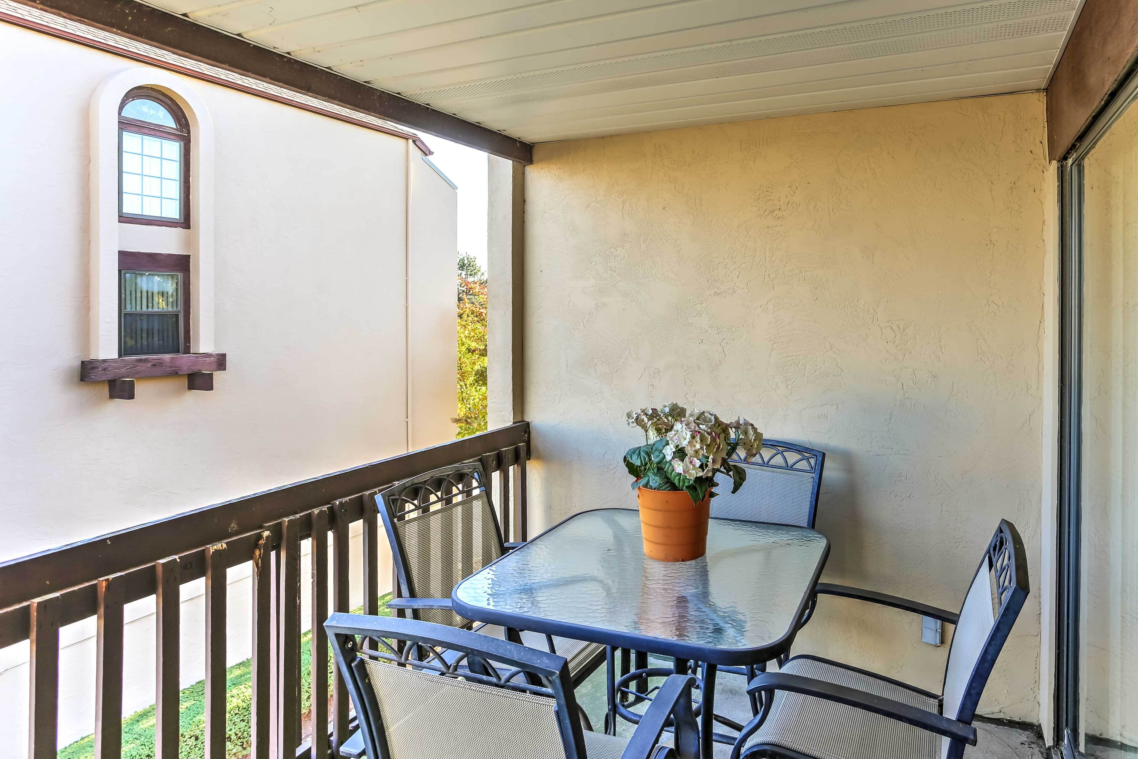 A balcony with a glass table and metal chairs, a potted plant, and a view of a building with a window and a door.