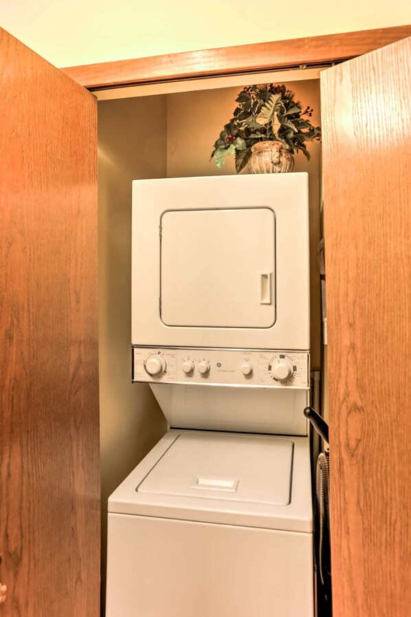 A compact laundry room with a washer and dryer, wooden cabinets, and a potted plant on top of the dryer. - Waterfront Port Clinton Condo w/ Pool Access! (Port Clinton)