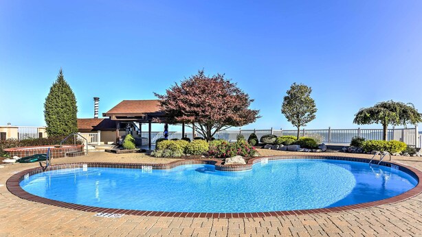 Une piscine avec une terrasse en briques, entourée d’arbres et bordée d’un bâtiment doté d’une cheminée.