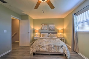 Une chambre à coucher avec un lit, deux tables de chevet avec des lampes, un ventilateur de plafond et une fenêtre avec des stores.