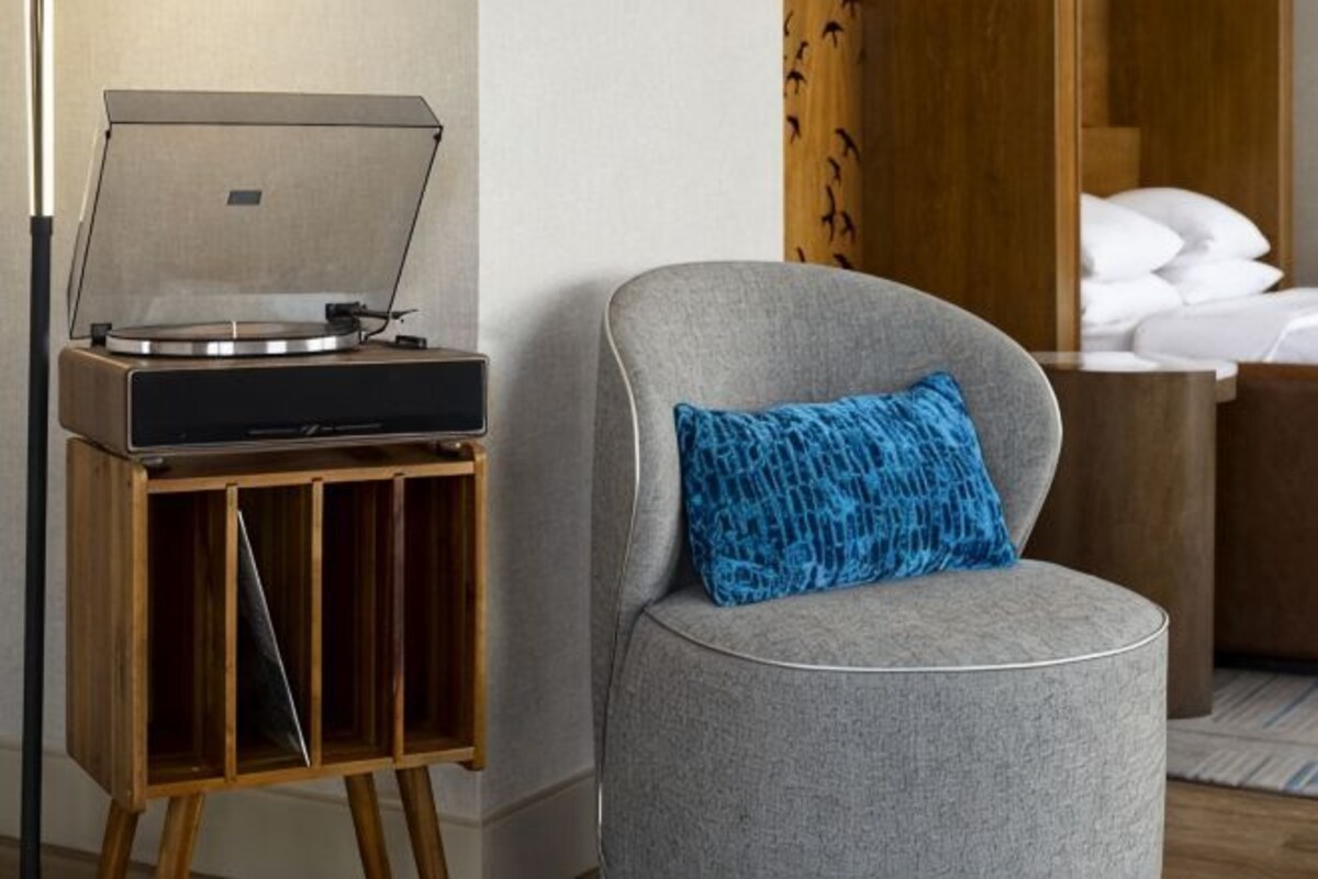 Een kamer met een grijze fauteuil, een houten bijzettafeltje met een platenspeler en een bed met witte lakens.