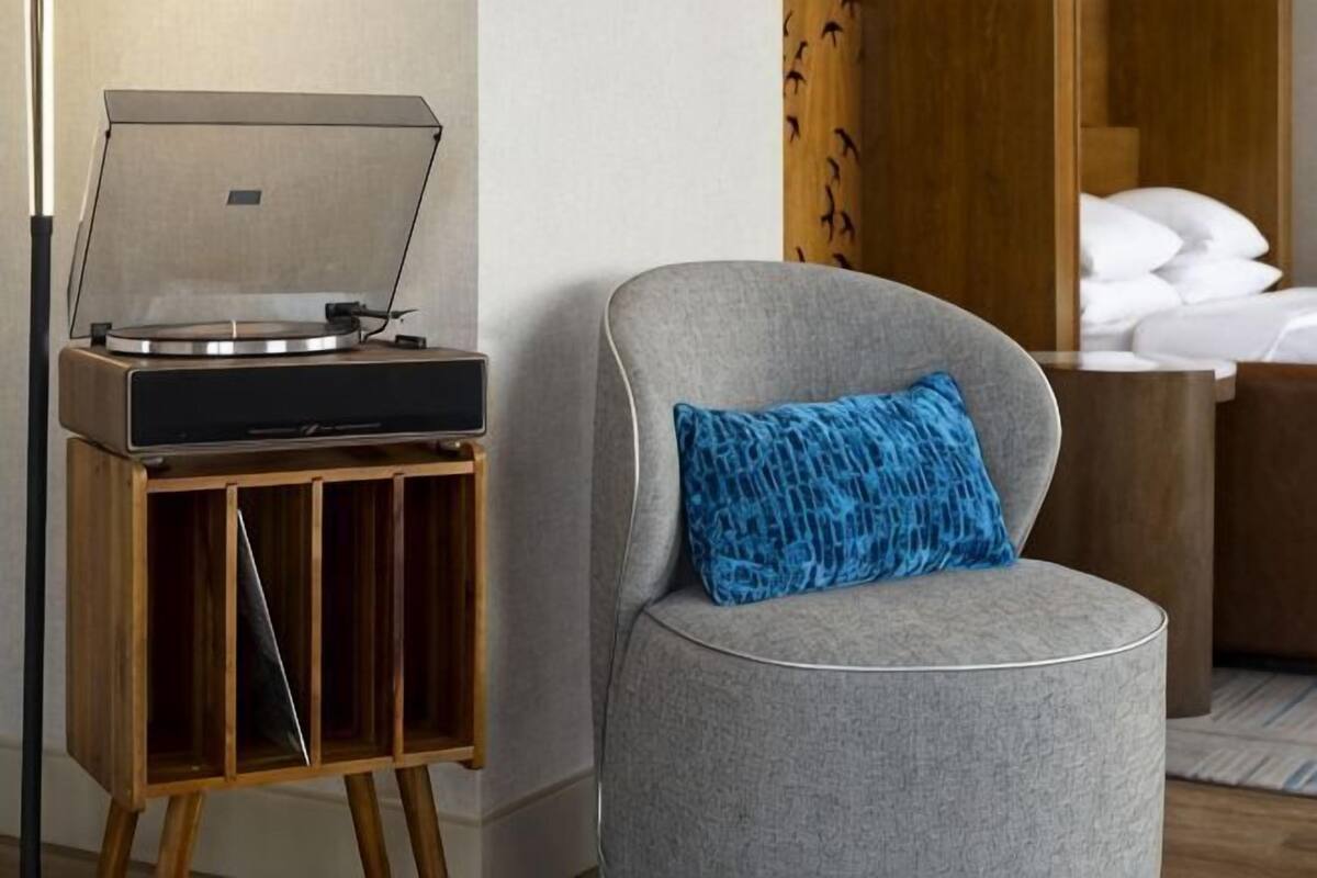 Une pièce avec un fauteuil gris, une table d’appoint en bois sur laquelle se trouve une platine vinyle, et un lit recouvert de draps blancs.