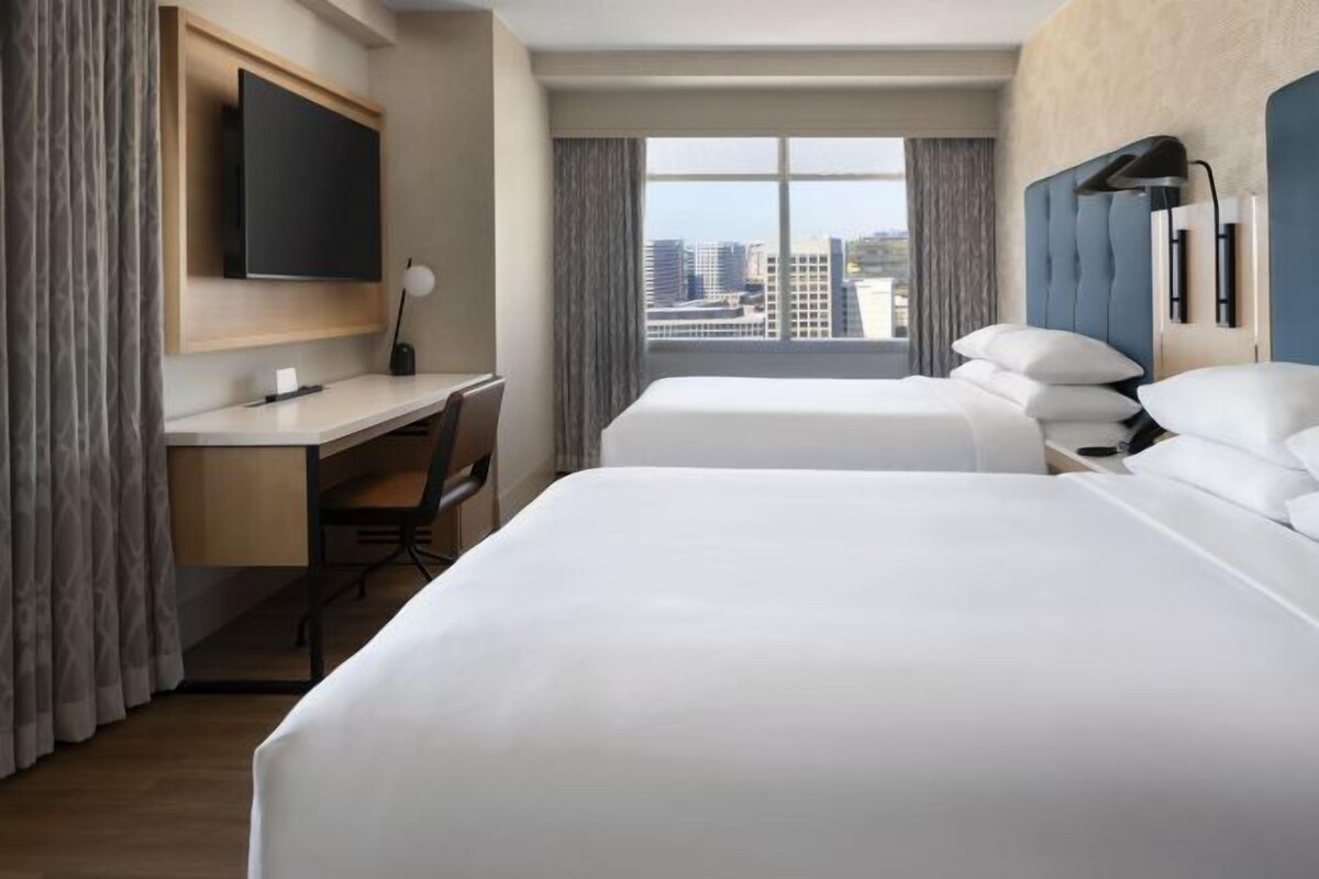 A hotel room with two beds, a desk, a TV, and a large window with a city view.