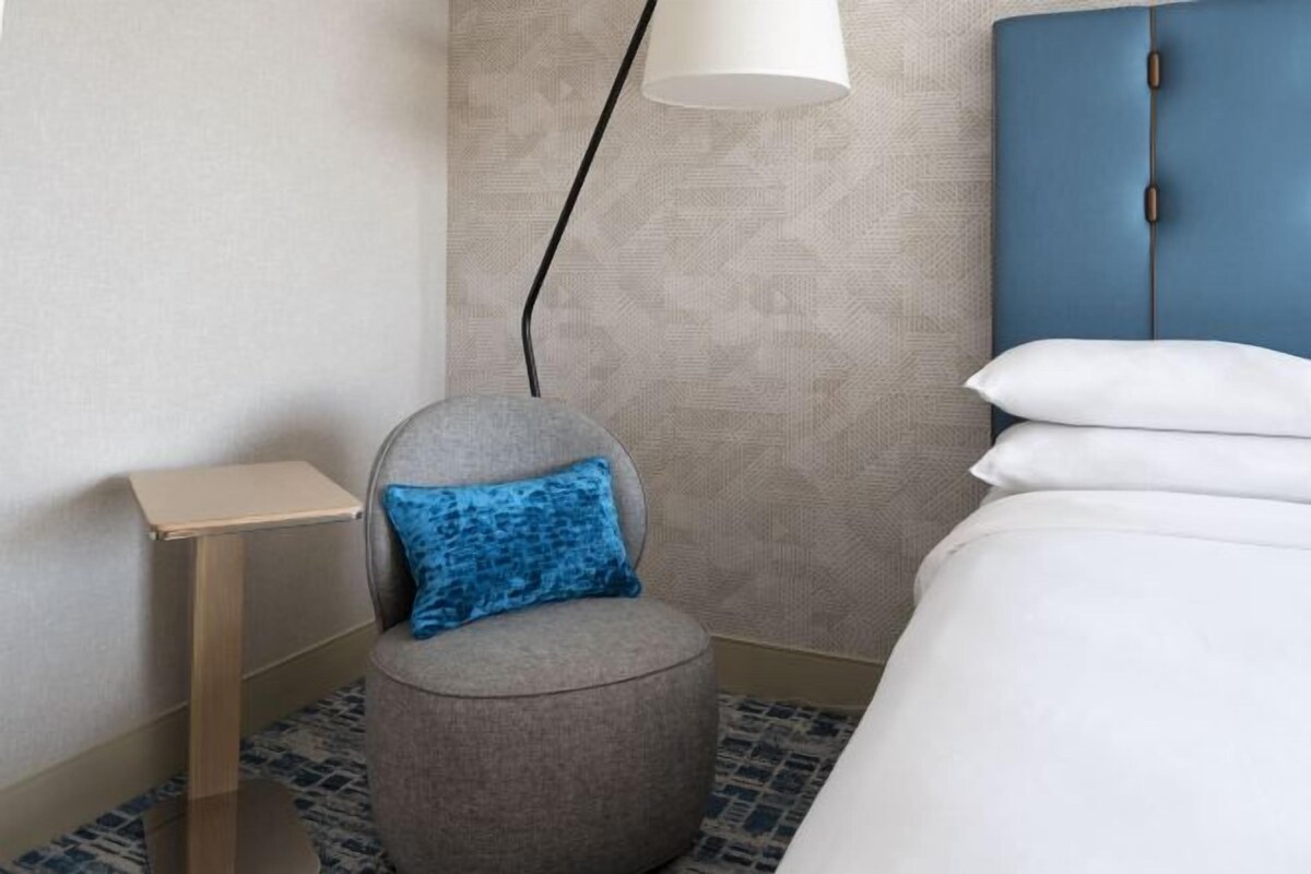 Et hotelværelse med en seng, en rund stol med blåt pudebetræk, et natbord og en lampe.