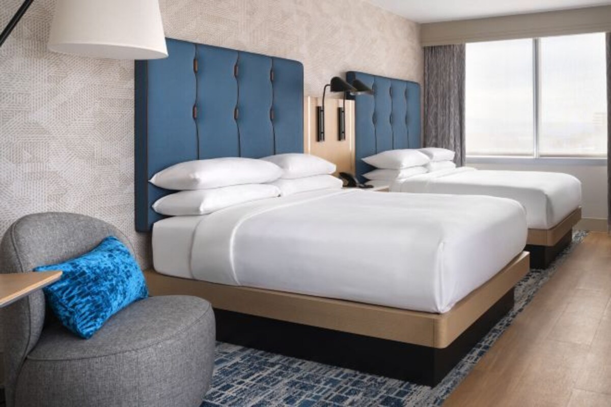 Een hotelkamer met twee bedden, een grijze fauteuil, een blauw kussen en een groot raam.