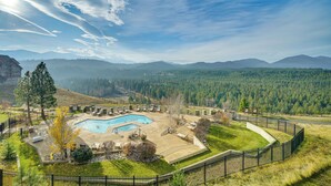 Una piscina circondata da un'area recintata con lettini, un edificio sullo sfondo e una catena montuosa boscosa.