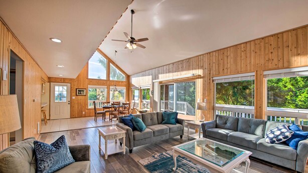A spacious living room with wooden paneling, a ceiling fan, and large windows offering views of the outdoors. - Hood Canal Home w/ Hot Tub - Bordering Olympic NP! (Brinnon)
