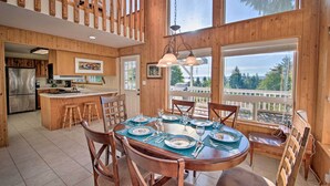 A wooden cabin interior with a dining area, kitchen, and large windows offering a view of the outdoors. - Hood Canal Home w/ Hot Tub - Bordering Olympic NP! (Brinnon)