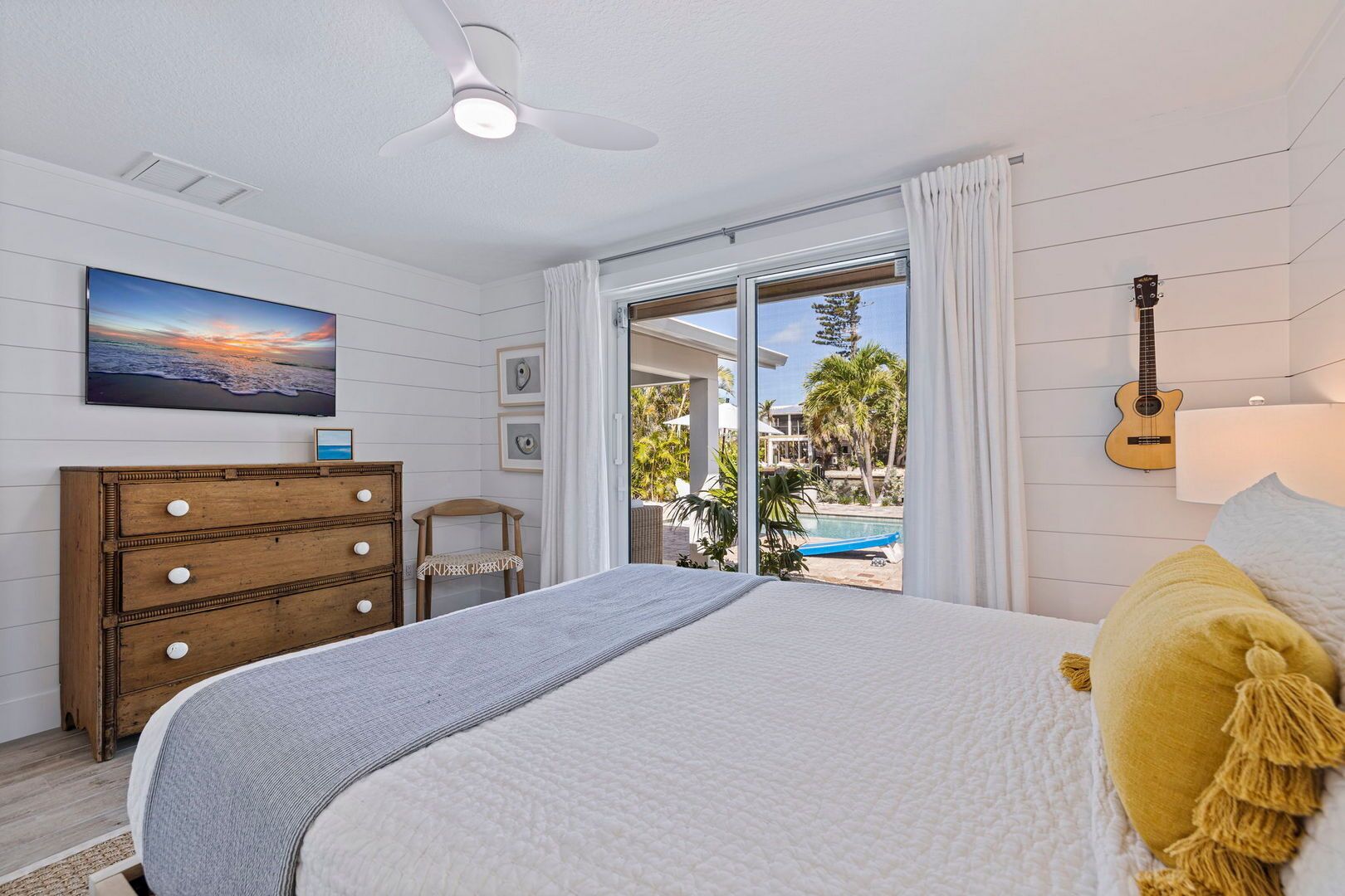 A bedroom with a bed, a wooden dresser, a ceiling fan, a TV, and a view of a pool and palm trees.