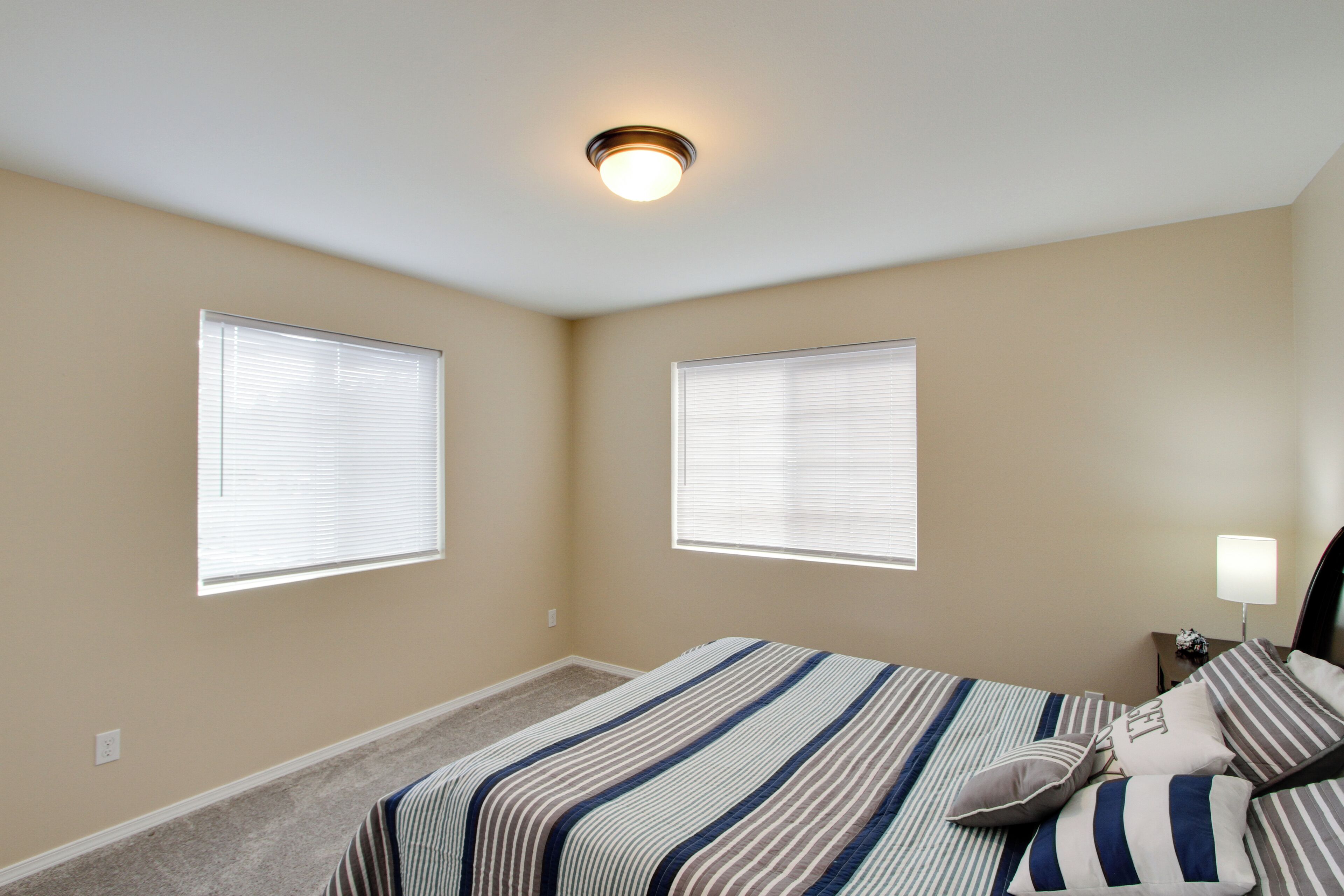 Un dormitorio con dos ventanas, una cama con ropa de cama a rayas, una mesita de noche con una lámpara y una luz en el techo.