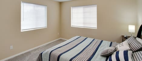 Un dormitorio con dos ventanas, una cama con ropa de cama a rayas, una mesita de noche con una lámpara y una luz en el techo.