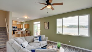 A spacious living room with a large window, a ceiling fan, and a kitchen area in the background. - Waterfront Allyn Home With Fire Pit + 2 Kayaks! (Allyn)