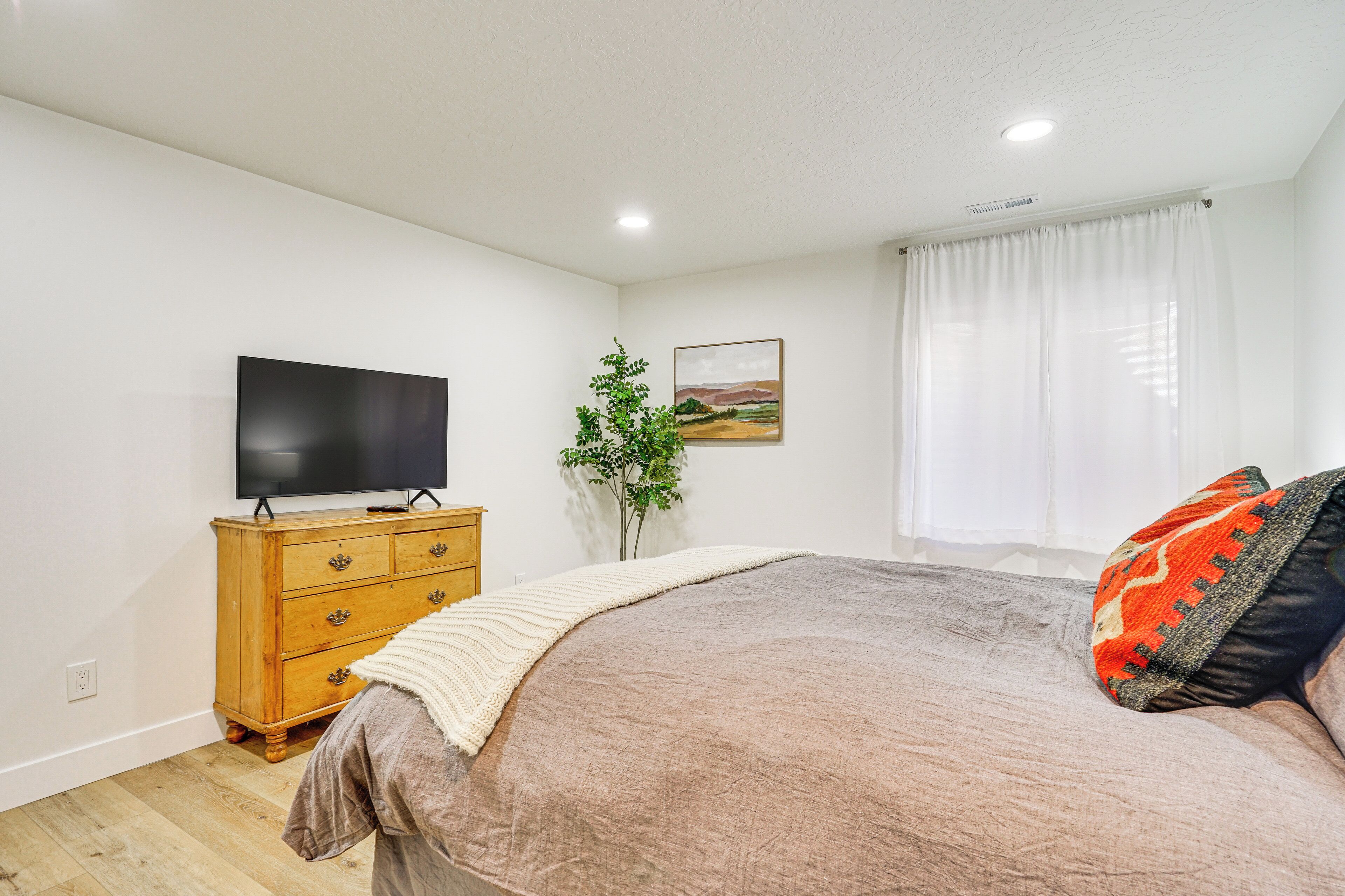 A bedroom with a bed, a TV on a wooden dresser, a plant, and a window with curtains.