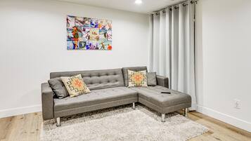 Un sofá seccional gris con cojines estampados, un alfombra de pelo alto blanco y un cartel colorido en la pared.
