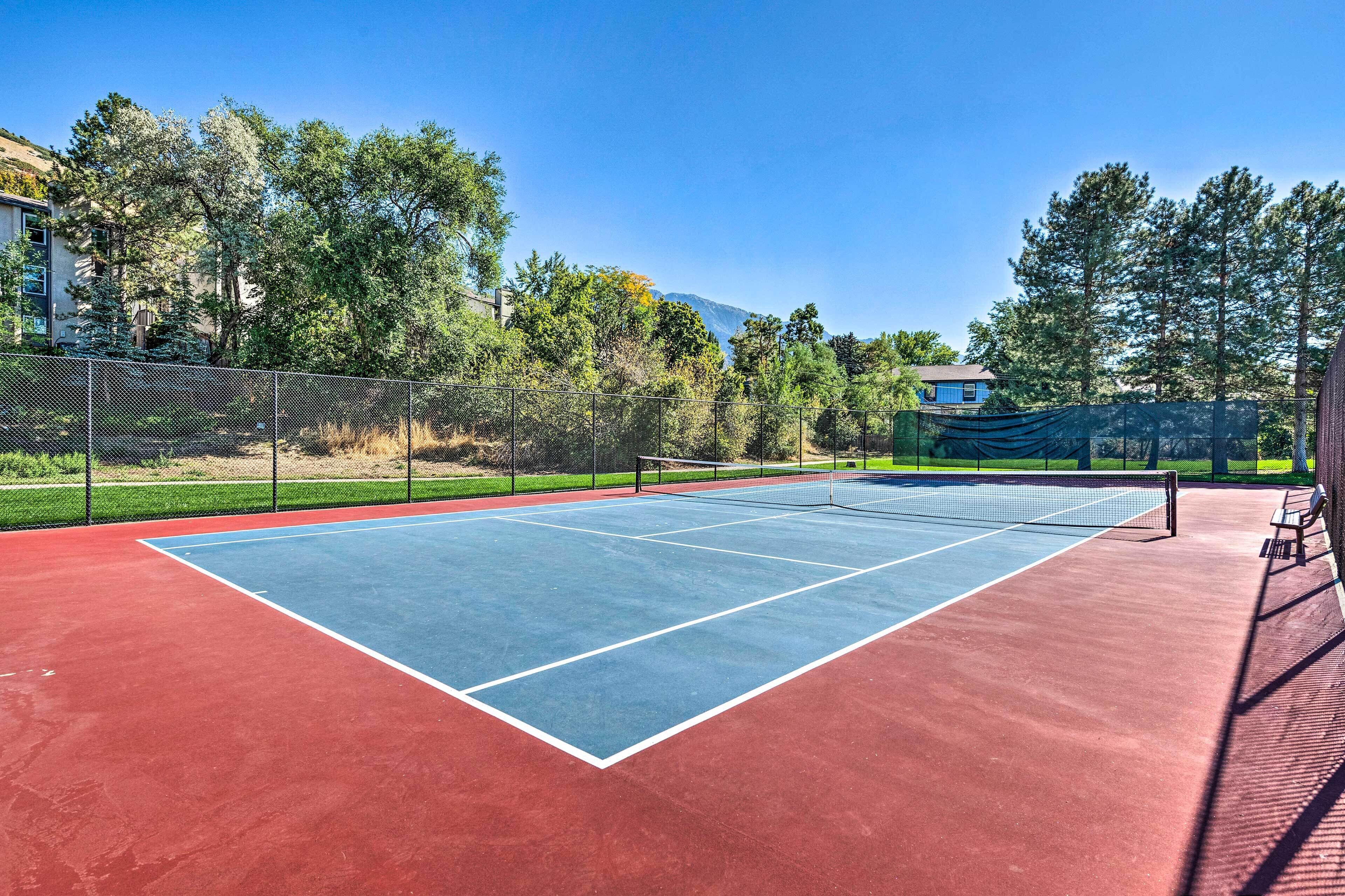 An outdoor tennis court with a net, surrounded by trees and a fence.