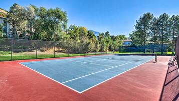 An outdoor tennis court with a net, surrounded by trees and a fence.