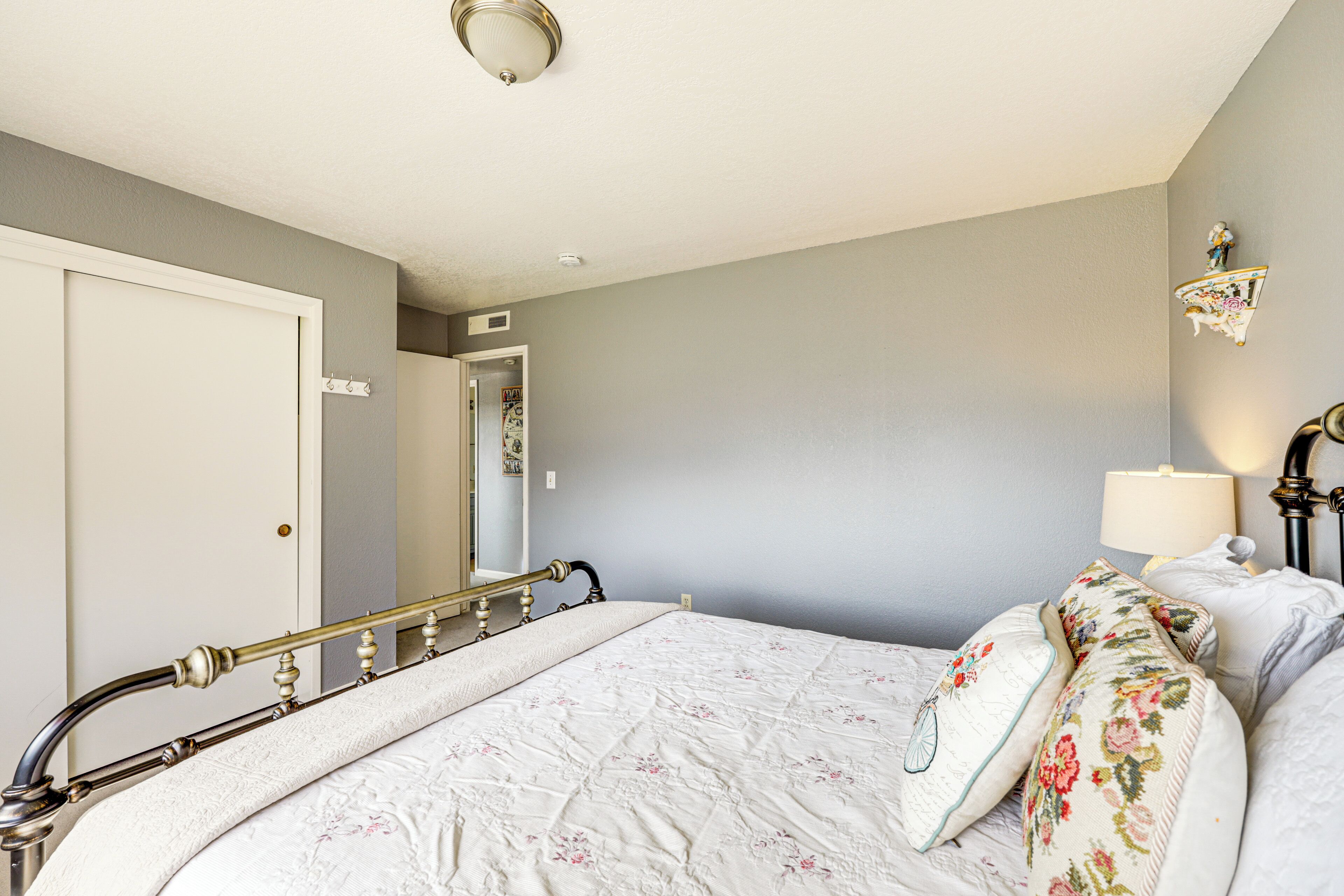 A neatly made bed with a floral comforter and pillows, a bedside table with a lamp, and a mirror on the wall.