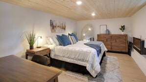 Une chambre avec un plafond en bois, un lit avec une literie à motifs, une commode, un bureau et une plante.