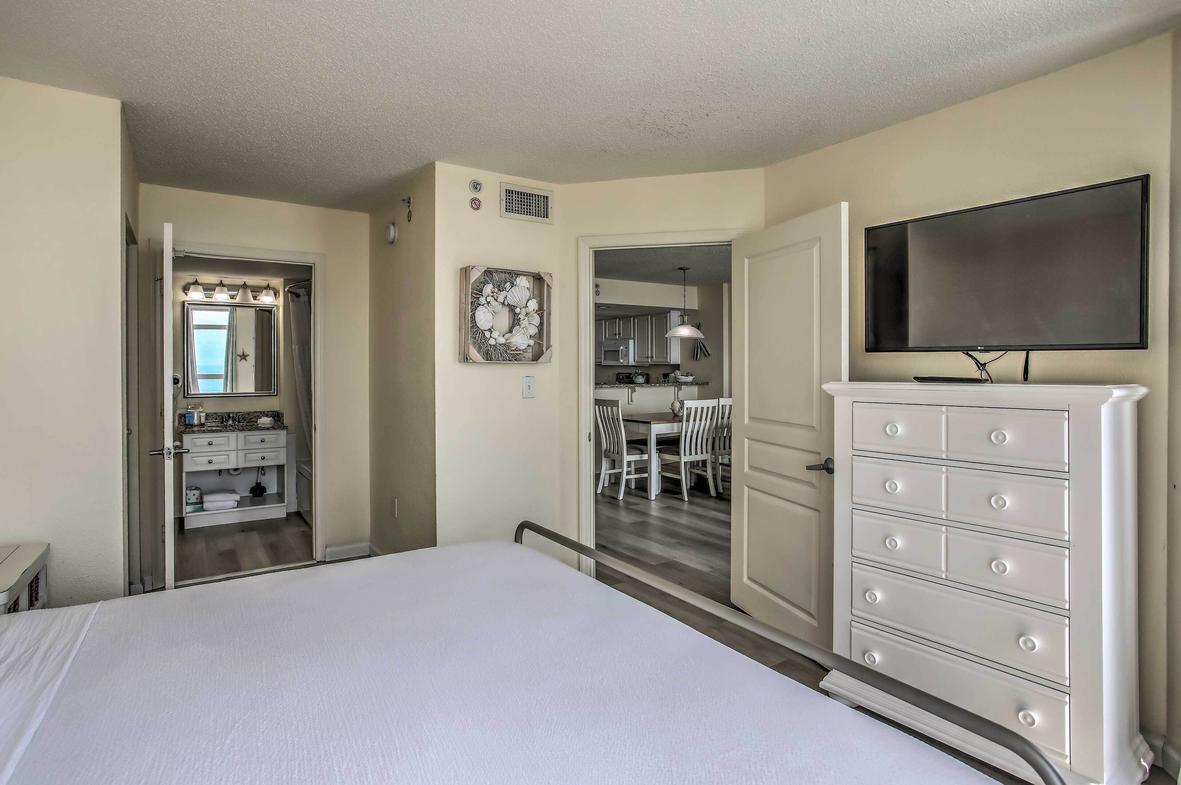 A hotel room with a bed, a dresser, a television, and a bathroom with a vanity and mirror.