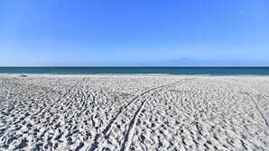 A wide sandy beach with tire tracks leading to the water, under a clear blue sky. - Bradenton Townhome - Walk to Locals-only Beach! (Bradenton Beach)