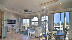 A spacious living room with a large TV, comfortable seating, and large windows offering a view of the outdoors. - Bradenton Townhome - Walk to Locals-only Beach! (Bradenton Beach)