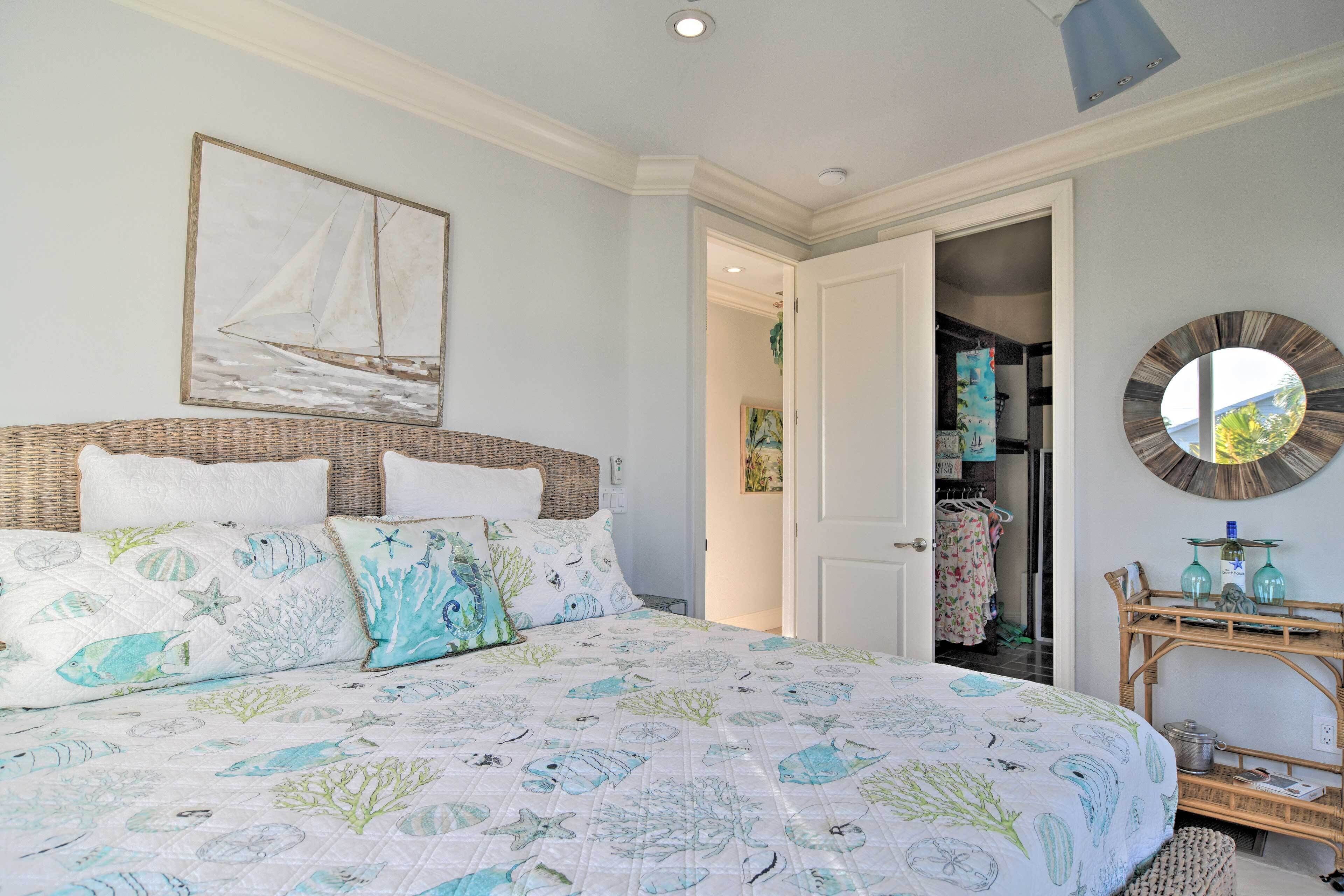 Een slaapkamer met een bed, een schilderij van een zeilboot aan de muur, een ronde spiegel en een rieten bijzettafeltje met glazen.