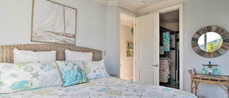 Een slaapkamer met een bed, een schilderij van een zeilboot aan de muur, een ronde spiegel en een rieten bijzettafeltje met glazen.