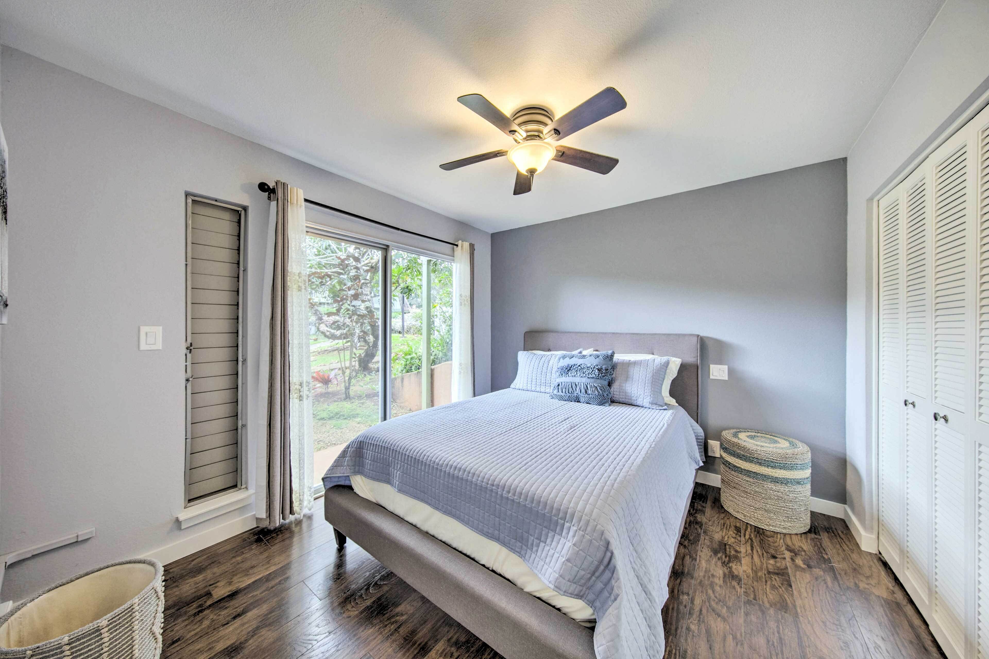 A bedroom with a bed, a ceiling fan, a window with curtains, and a small round ottoman.