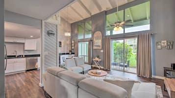 A modern living room with a large sofa, a coffee table, and a dining area with a table and chairs. There is a kitchen with white cabinets and a dishwasher. The room has large windows with curtains and a view of the outdoors.