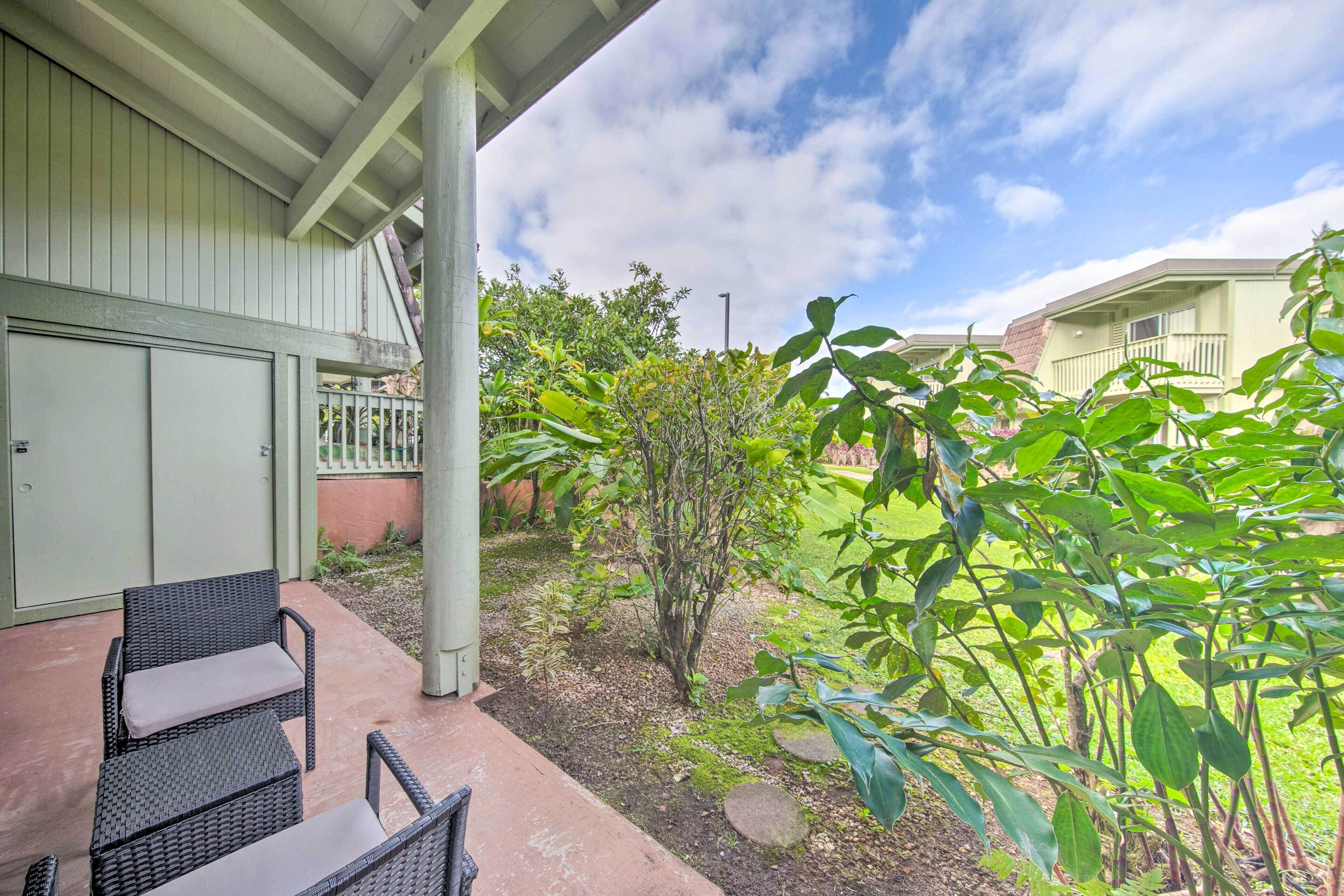 A covered patio with wicker furniture, a small garden area with various plants, and a view of neighboring buildings.