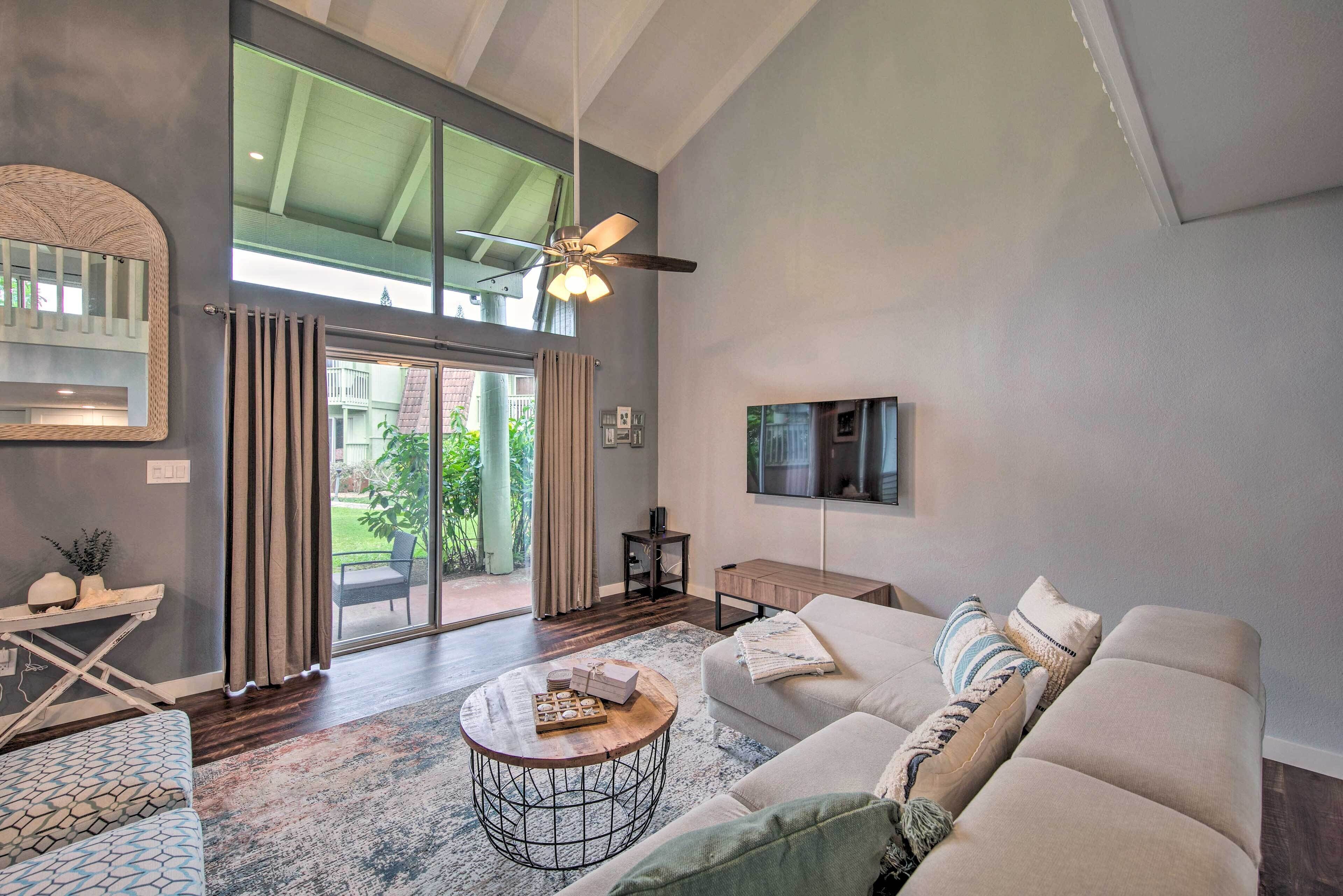 A spacious living room with a large window, a ceiling fan, a sofa, a coffee table, and a TV mounted on the wall.
