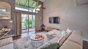 A spacious living room with a large window, a ceiling fan, a sofa, a coffee table, and a TV mounted on the wall. - Coastal Escape Condo w/ Pool, Near Beaches! (Princeville)