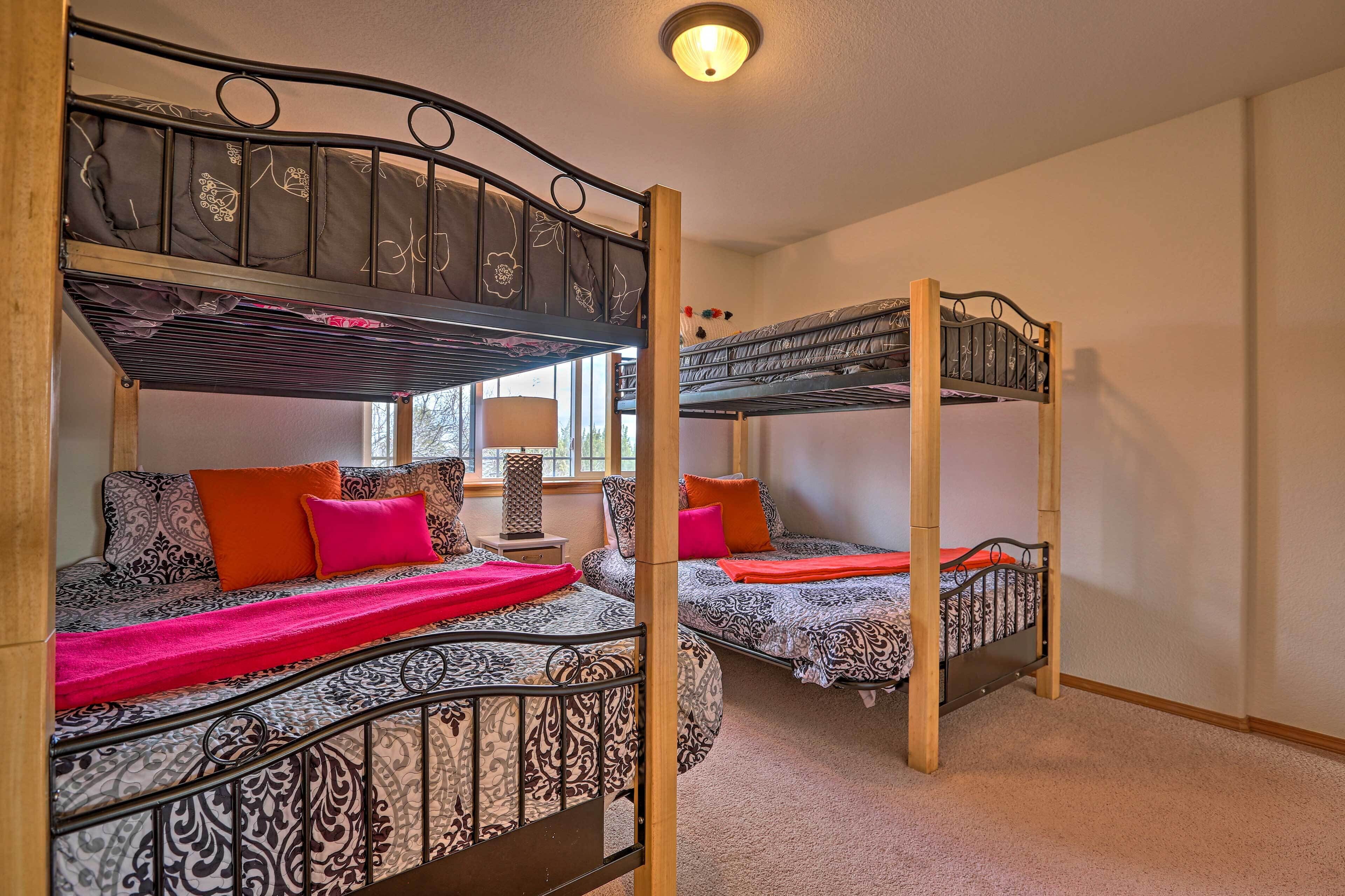 A room with two bunk beds, each with patterned bedding and pillows, a bedside table with a lamp, and a window providing natural light.