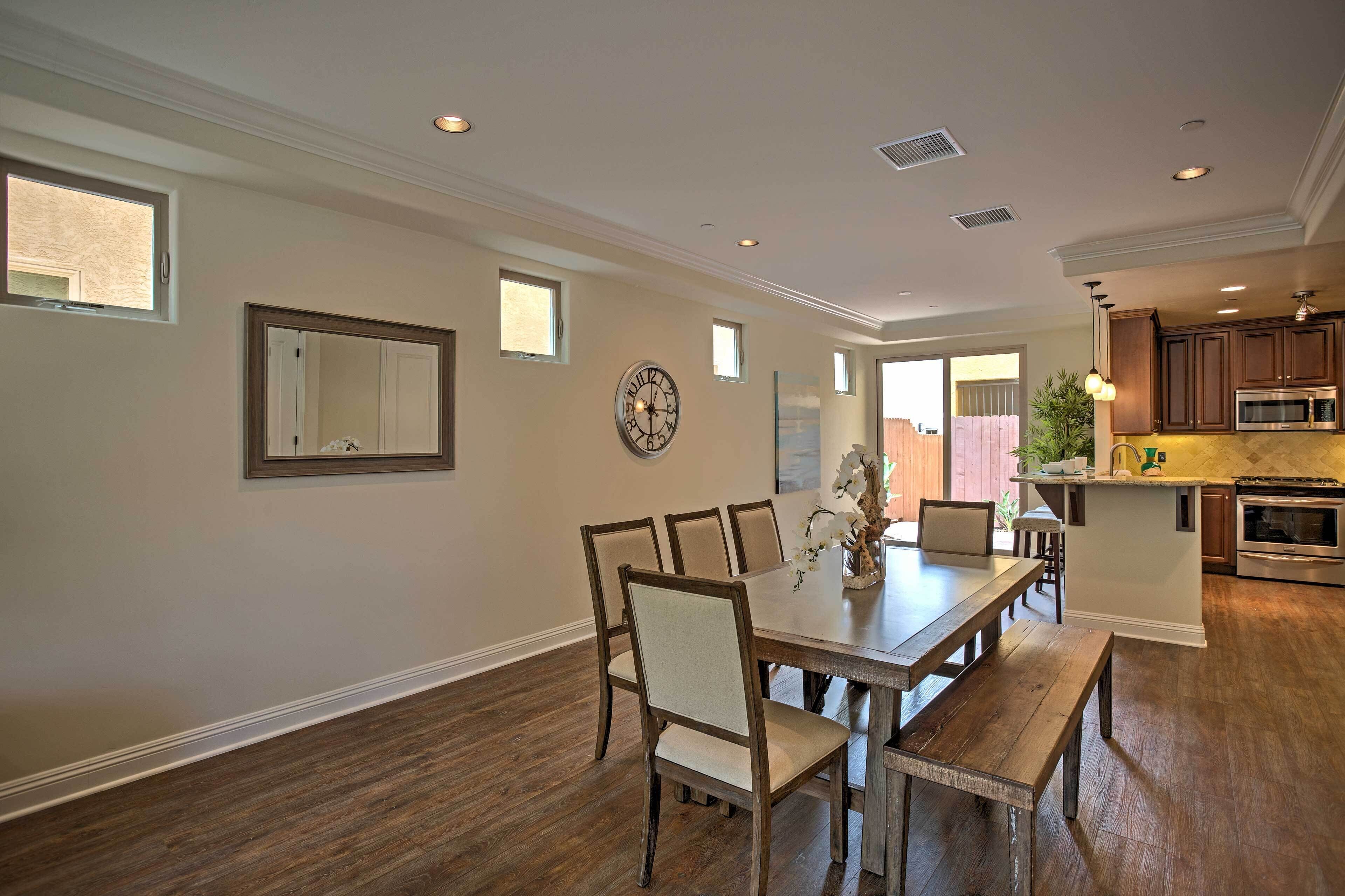 A dining area with a wooden table and chairs, a clock on the wall, and a kitchen in the background.