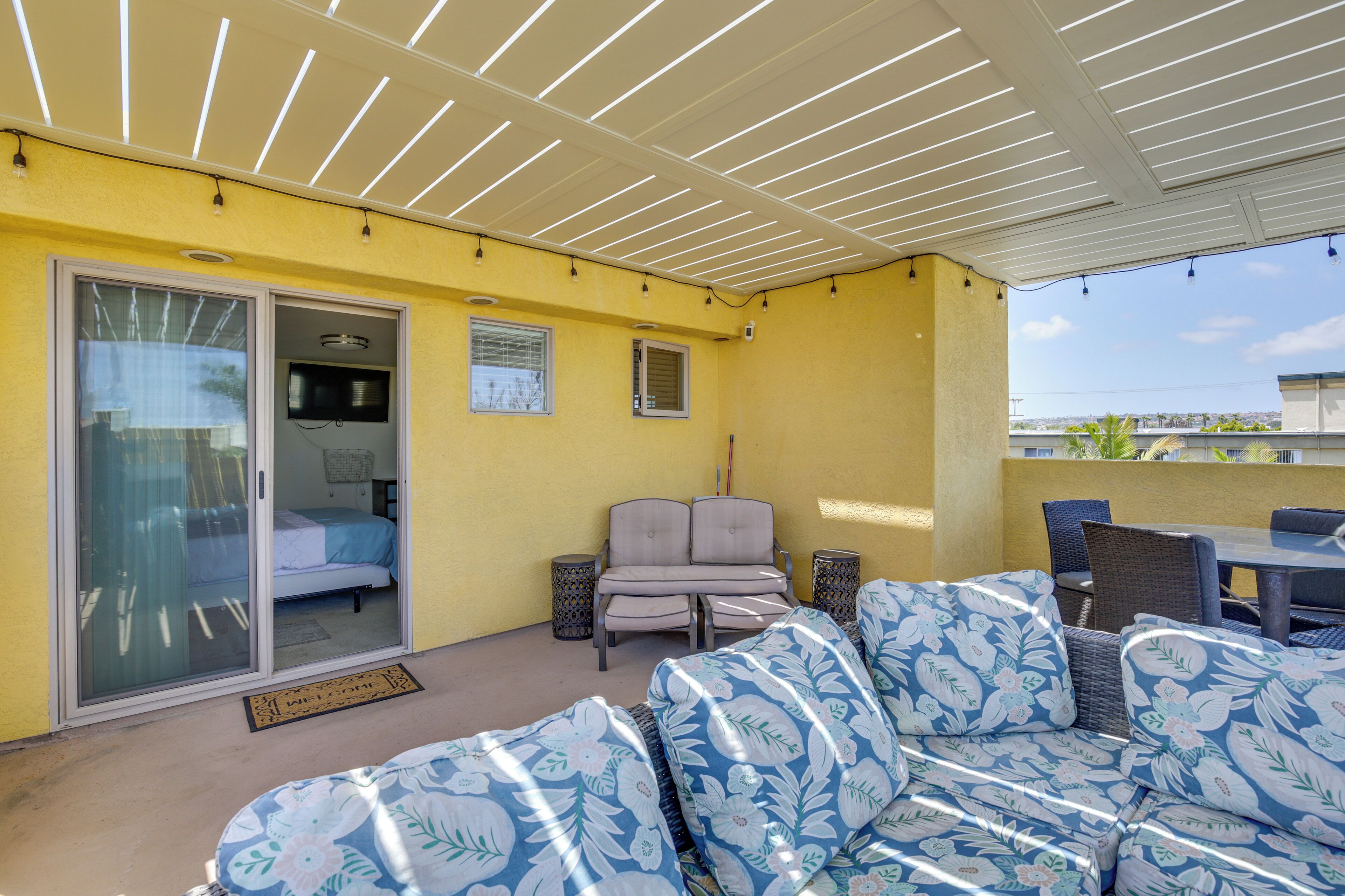 A covered patio area with a yellow wall, a sliding glass door leading to a bedroom, a sofa set, and a table with chairs.