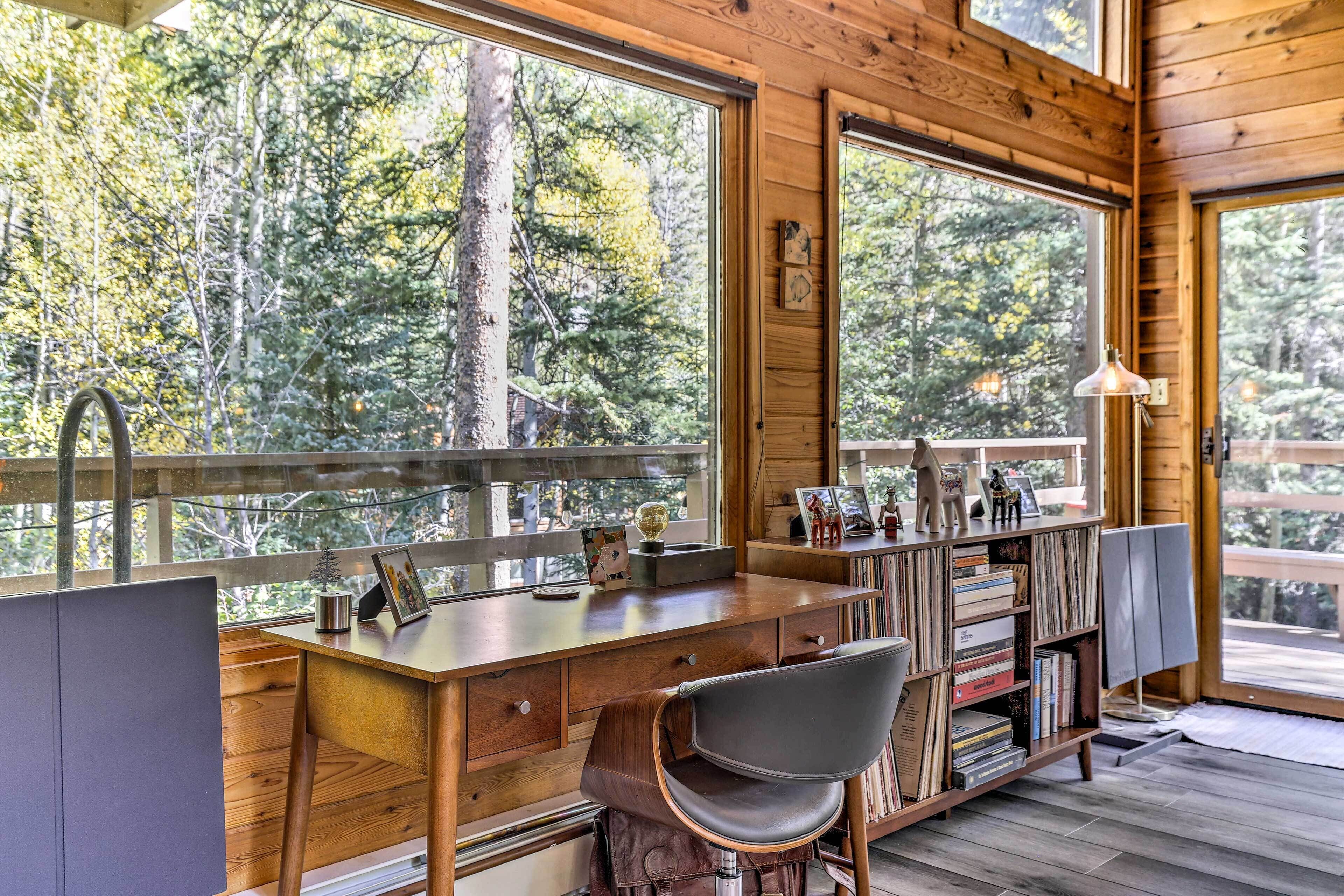 Interior de una cabaña de madera con escritorio, silla, estantería y ventanas amplias con vistas a un bosque.