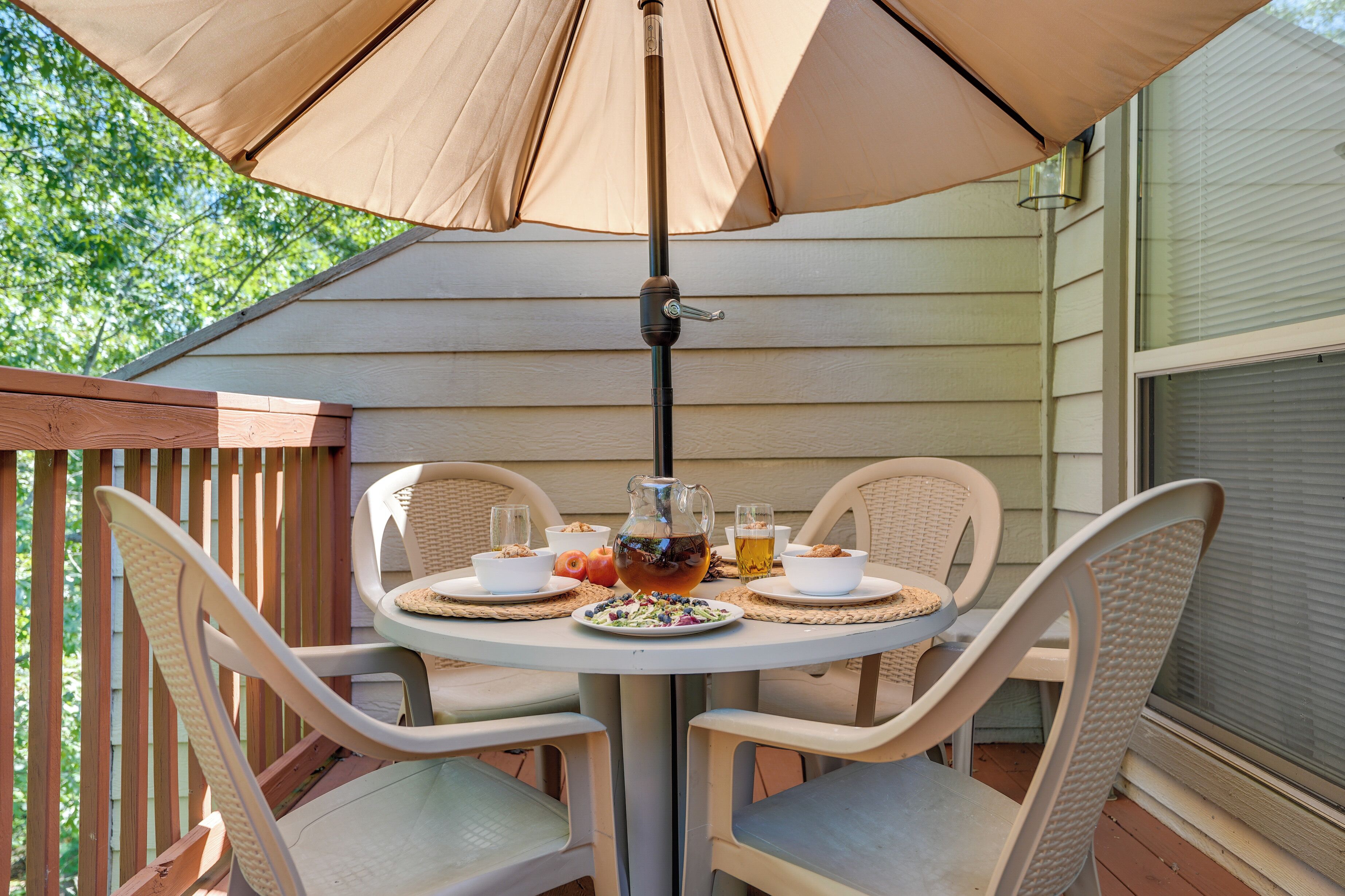 A round table with four chairs, a pitcher, bowls, and glasses, under a large umbrella on a wooden deck.