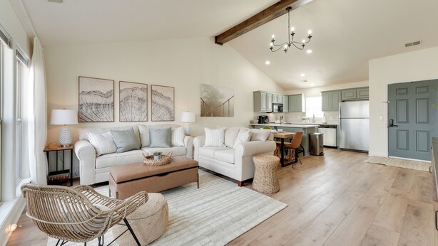 Un espacio habitable amplio con un conjunto de sofás, una mesa de centro y una silla de mimbre. Se ve una cocina con electrodomésticos modernos al fondo.