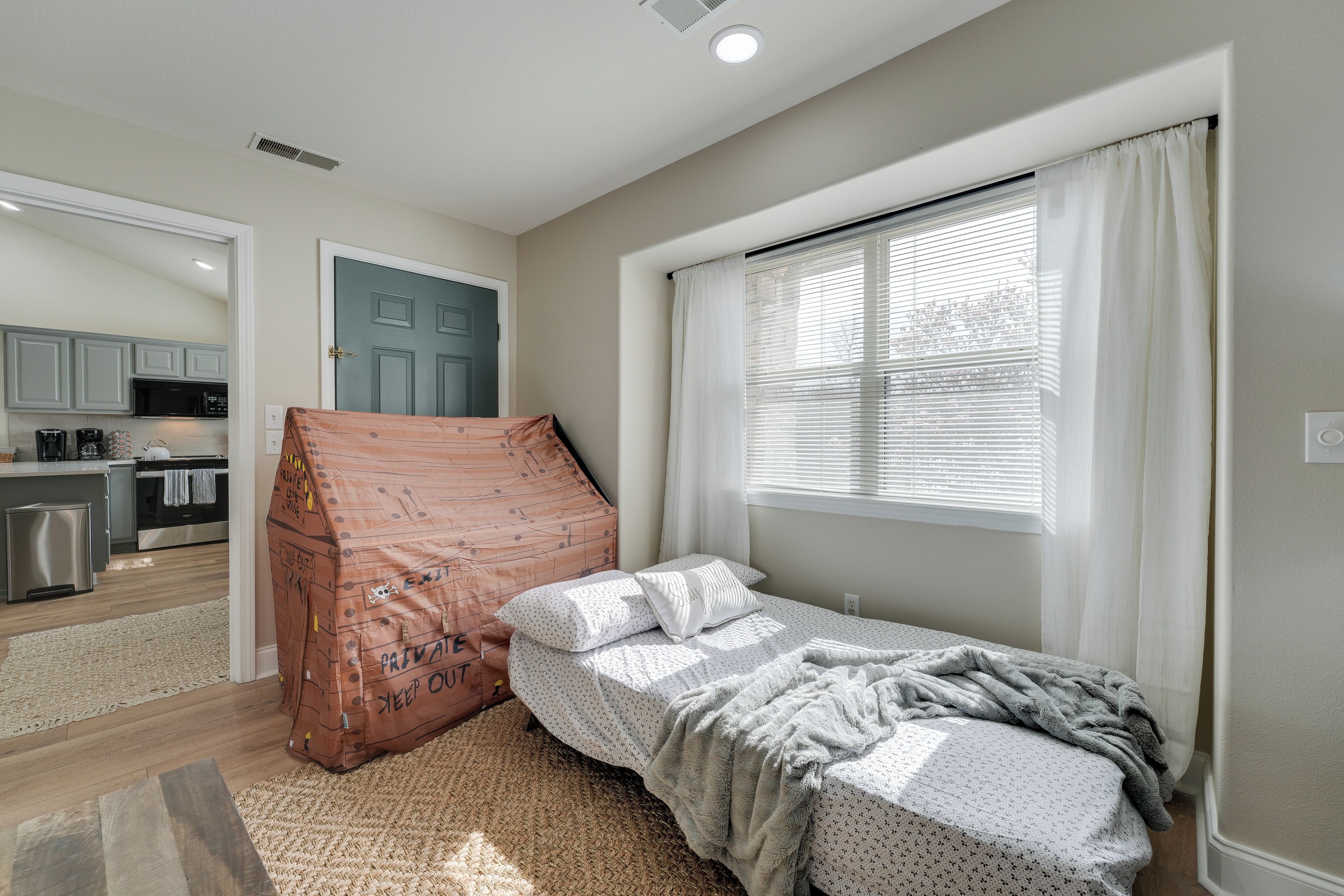 A bedroom with a bed, a window with curtains, a kitchenette in the background, and a door.