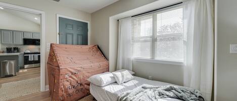 A bedroom with a bed, a window with curtains, a kitchenette in the background, and a door.