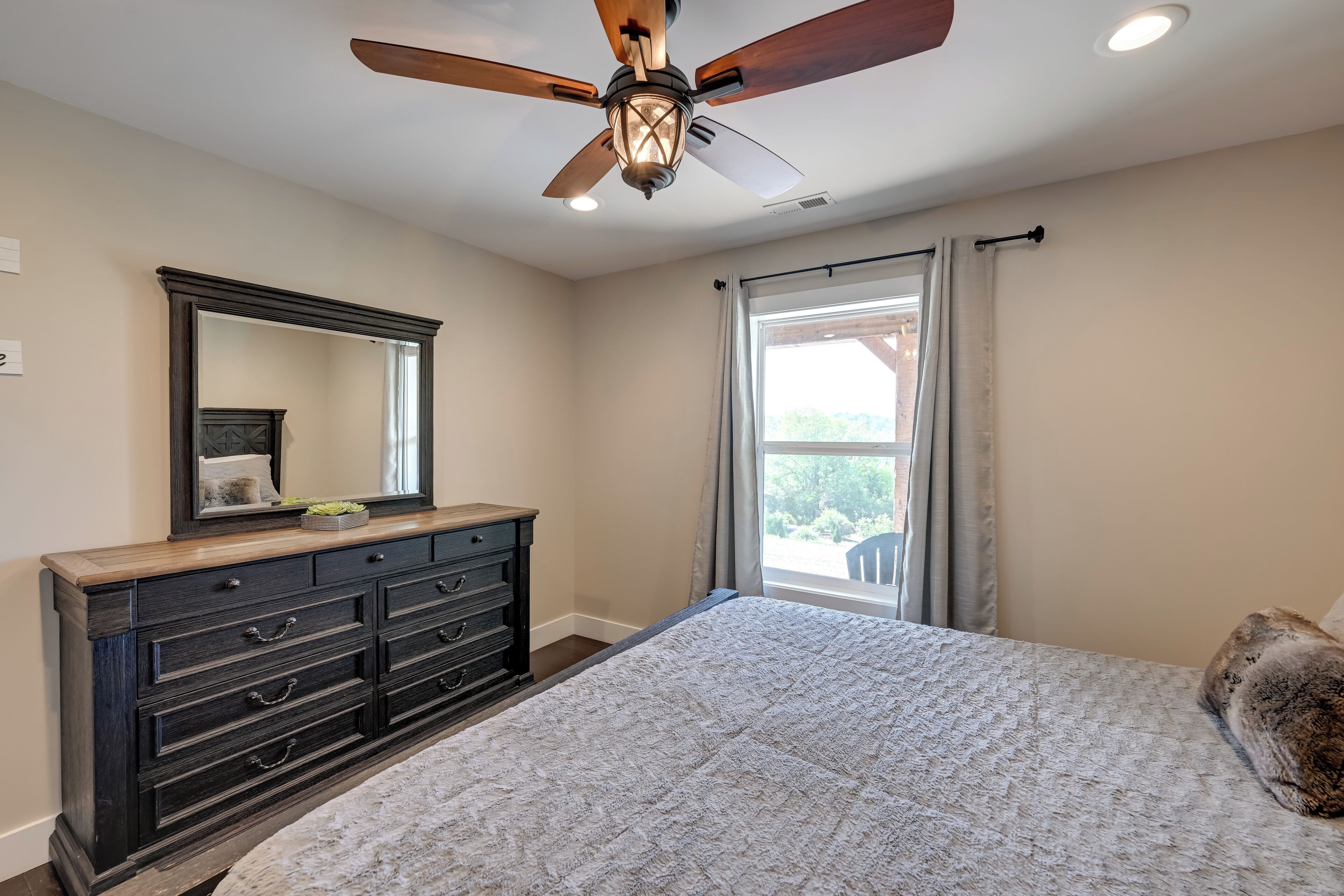 A bedroom with a large bed, a dark wooden dresser, a ceiling fan, and a window with curtains.