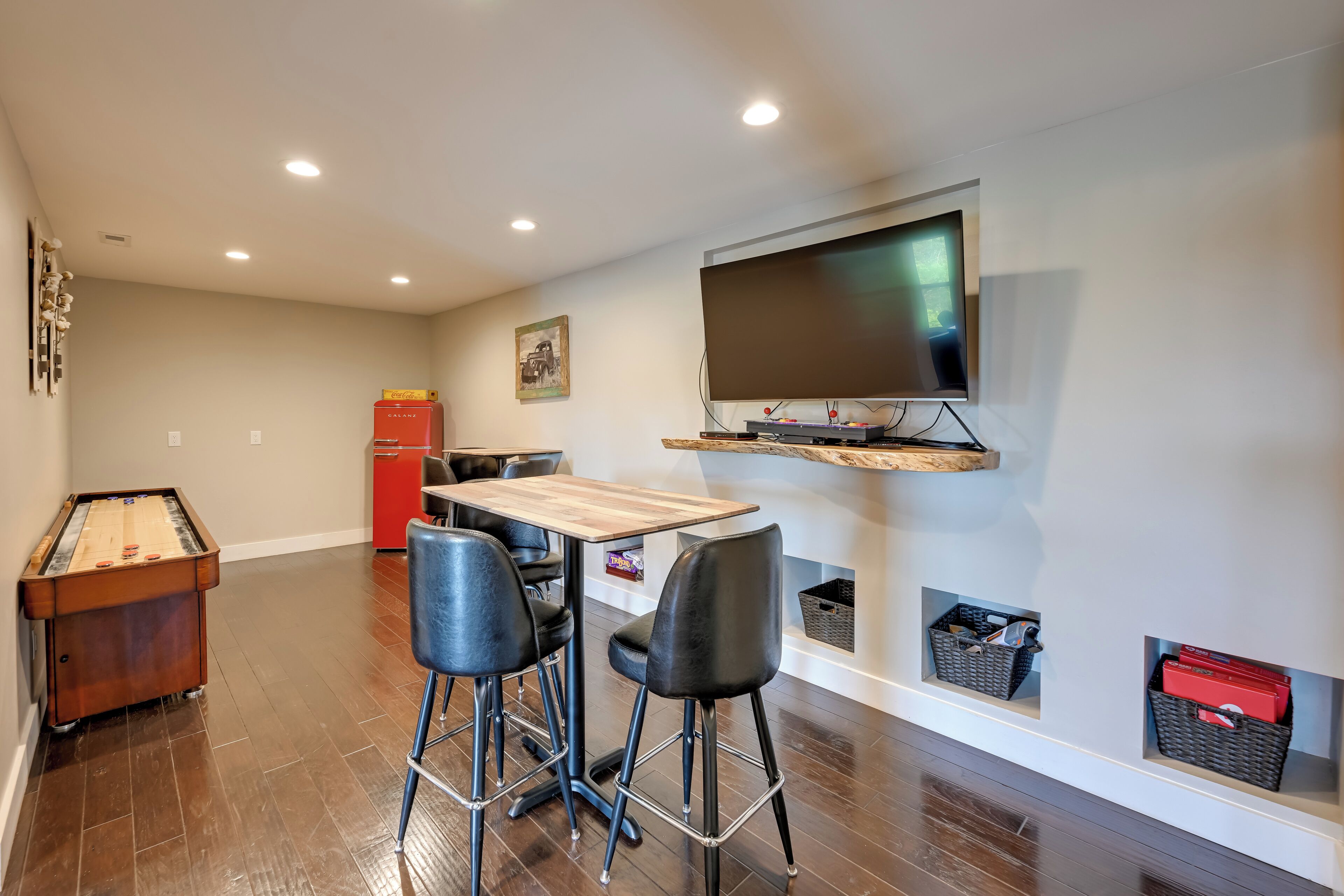 A living room with a pool table, two bar stools, a wooden table, a flat-screen TV mounted on the wall, and storage baskets under the TV.