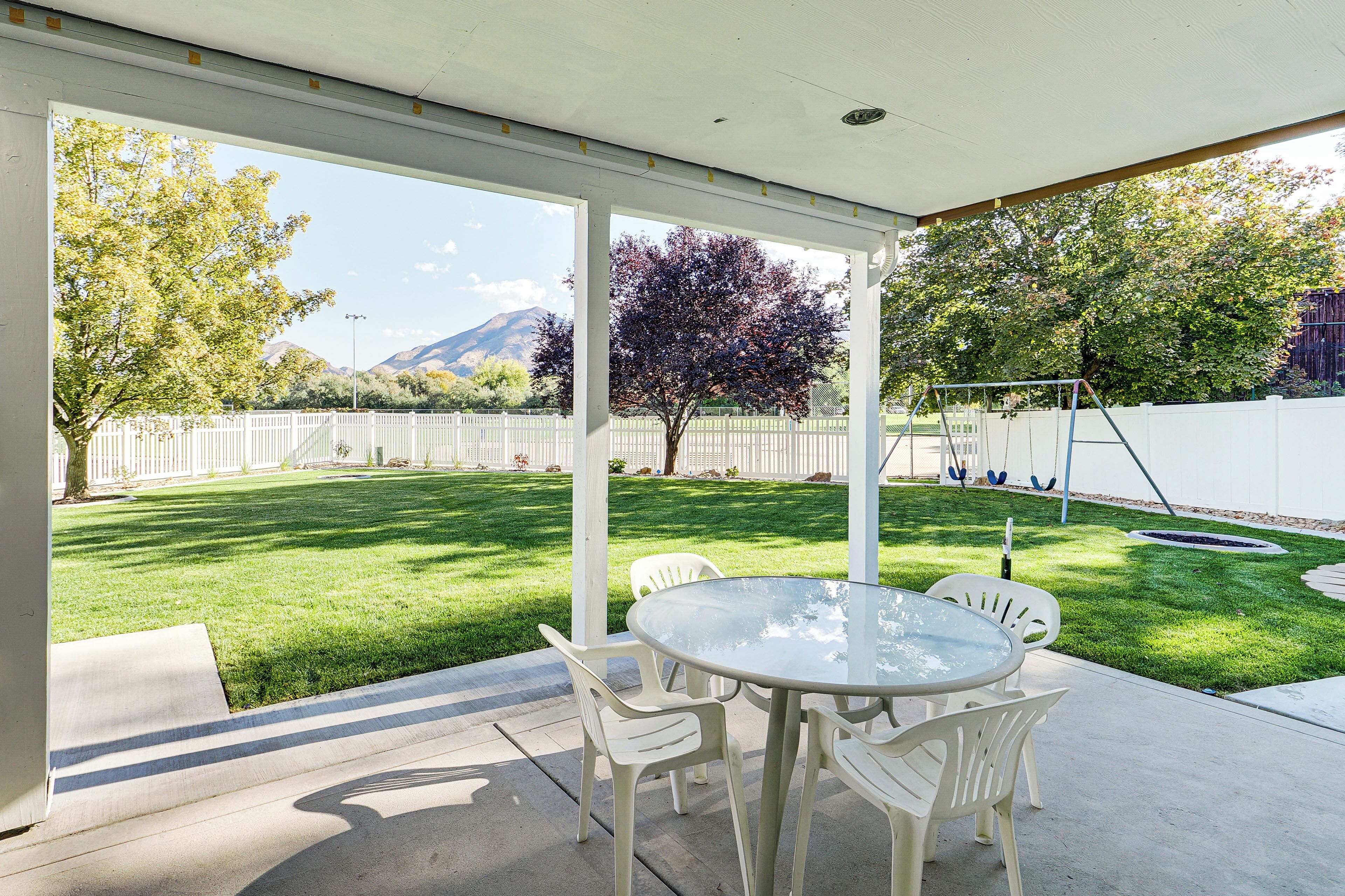 A covered patio with a table and chairs, a lawn, and a white fence.