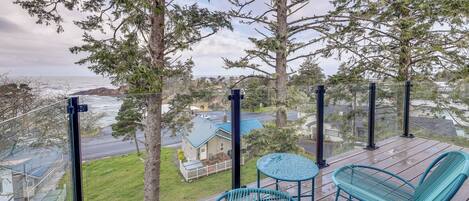 Um deck de madeira com duas cadeiras e uma mesa pequena, com vista para uma área residencial e o oceano.