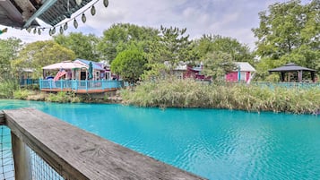 Eine Holzterrasse mit Blick auf ein türkisblaues Schwimmbecken, mit überdachter Sitzecke und einem Pavillon im Hintergrund.