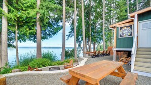 A wooden picnic table with benches, a fire pit, and a building with a sign in the background. - Fish, Crab & Clam: Waterfront Gem in Port Ludlow (Port Ludlow)