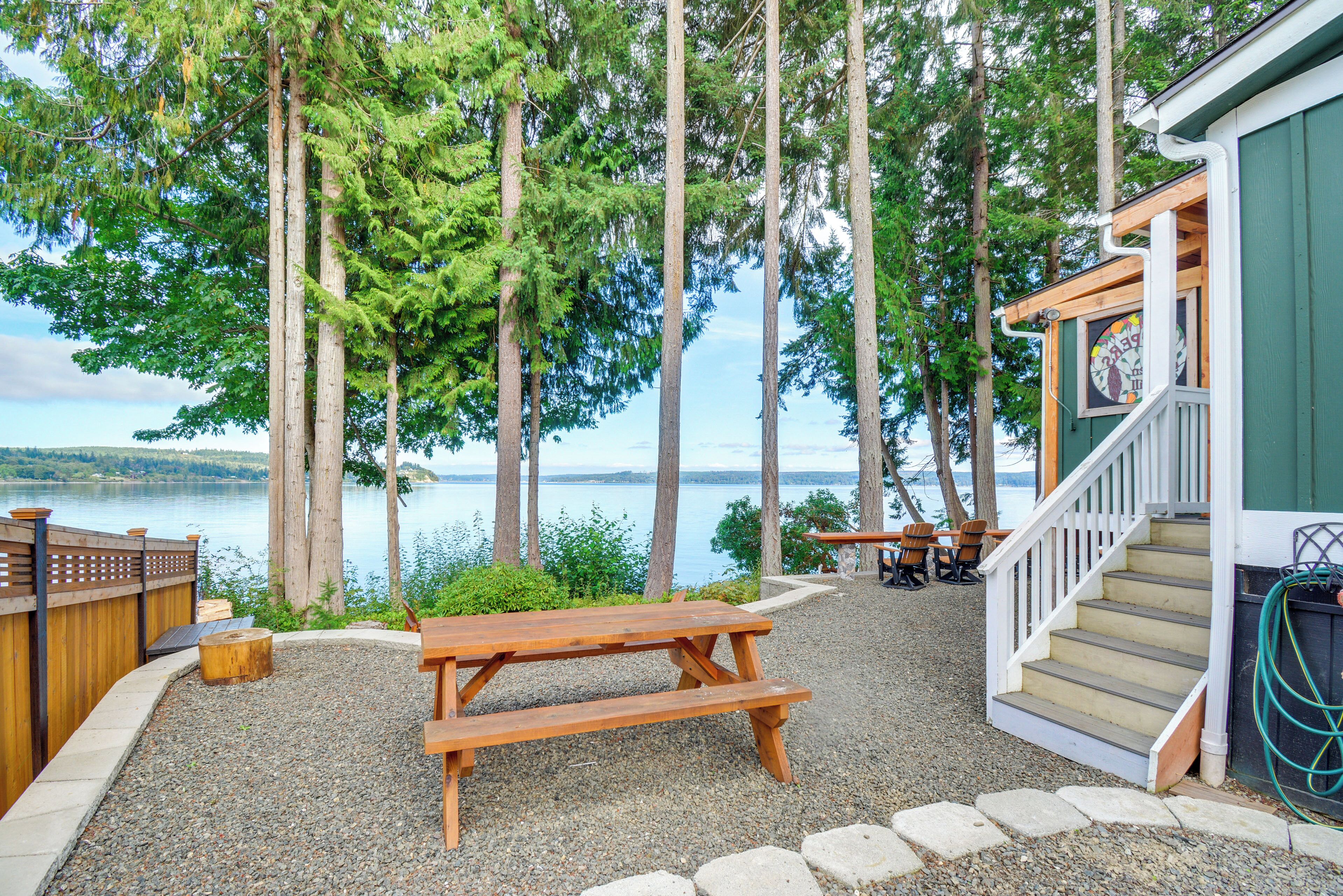 A wooden picnic table and benches, a staircase leading to a building, and a view of a lake surrounded by tall trees.