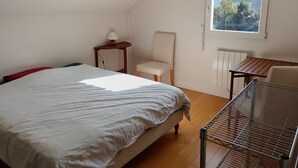 A bedroom with a bed, a chair, a small table, a radiator, and a window with a view of buildings and trees. - Homestay (Chambéry)